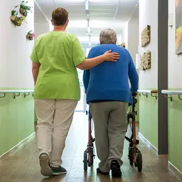 Eine Pflegefachkraft geht mit einer Seniorin den Flur einer Pflegeeinrichtung entlang.